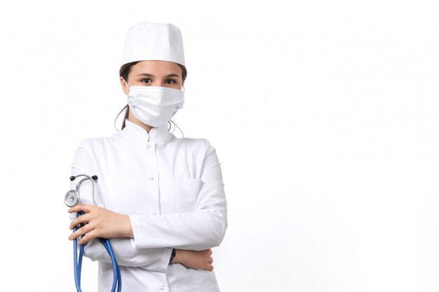 A front view young female doctor in white medical suit and white cap with blue stethoscope wearing a mask 