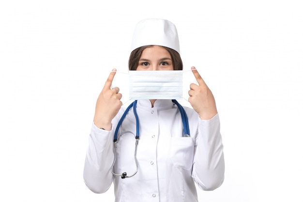 A front view young female doctor in white medical suit and white cap with blue stethoscope wearing a mask 