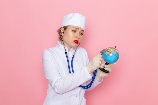 Giovane medico femminile di vista frontale in vestito medico bianco con lo stetoscopio blu che controlla il piccolo globo sul lavoro medico dell'ospedale della medicina dello spazio rosa