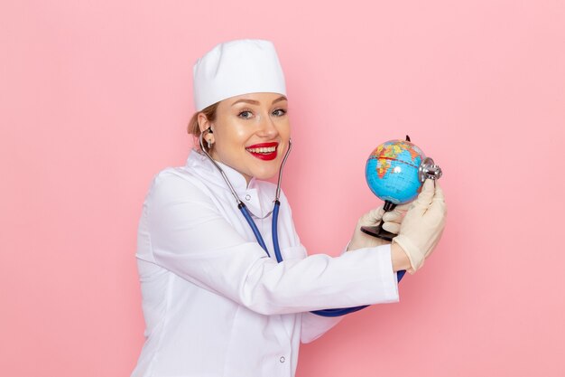 Вид спереди молодая женщина-врач в белом медицинском костюме с синим стетоскопом проверяет маленький глобус, улыбаясь в розовой медицинской больнице космической медицины