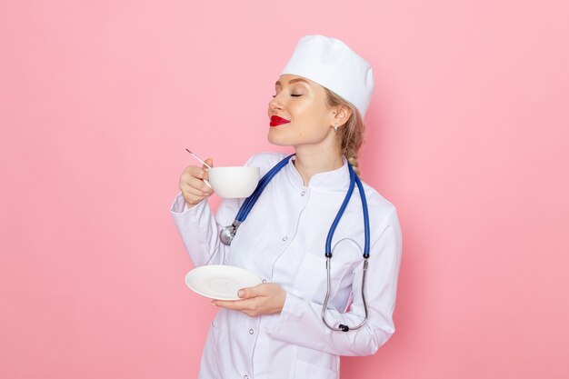 Front view young female doctor in white medical suit with blue stethoscope drinking coffee on the pink space medicine medical hospital job