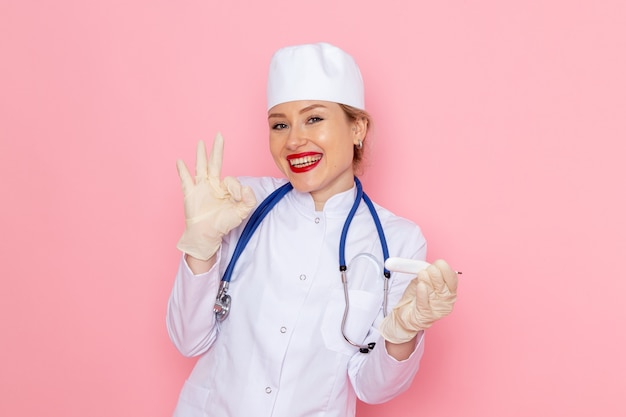 Вид спереди молодая женщина-врач в белом медицинском костюме с синим стетоскопом, держащим устройство, улыбаясь в розовой медицинской больнице космической медицины