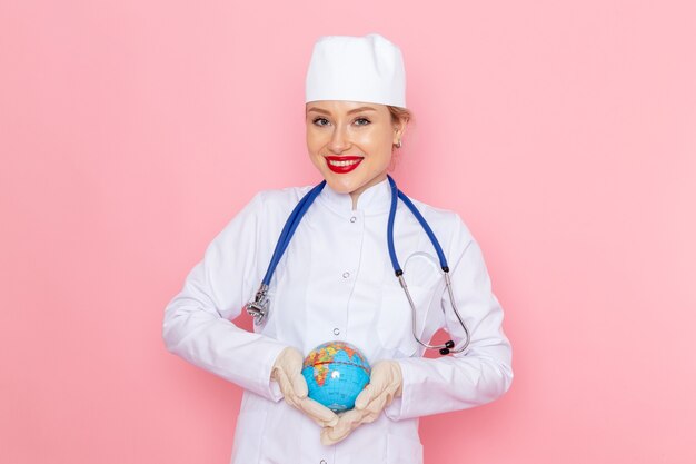 Front view young female doctor in white medical suit with blue stethoscope holding globe with smile on the pink space medicine medical hospital health