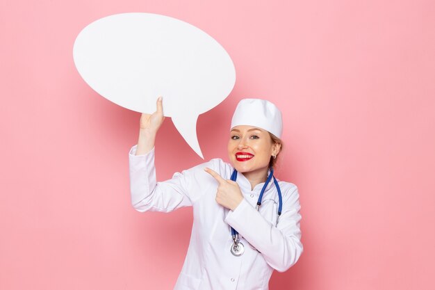 Front view young female doctor in white medical suit with blue stethoscope holding huge white sign smiling on the pink space