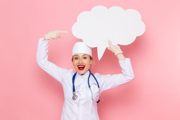 Вид спереди молодая женщина-врач в белом медицинском костюме с синим стетоскопом, держащая огромный белый знак с улыбкой на розовом медицинском госпитале космической медицины