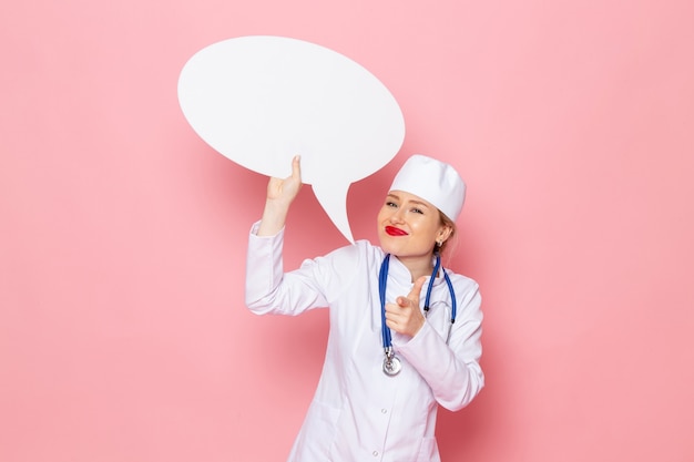 Front view young female doctor in white medical suit with blue stethoscope holding huge white sign with smile on the pink space