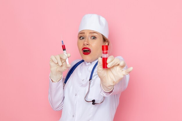 Вид спереди молодая женщина-врач в белом медицинском костюме с синим стетоскопом, держащая инъекцию и колбу на розовом космическом медицинском здоровье