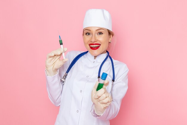 Front view young female doctor in white medical suit with blue stethoscope holding injection and flask smiling on the pink floor medicine medical hospital health