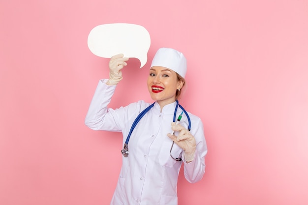 Front view young female doctor in white medical suit with blue stethoscope holding injection and white sign smiling on the pink space medicine medical hospital job