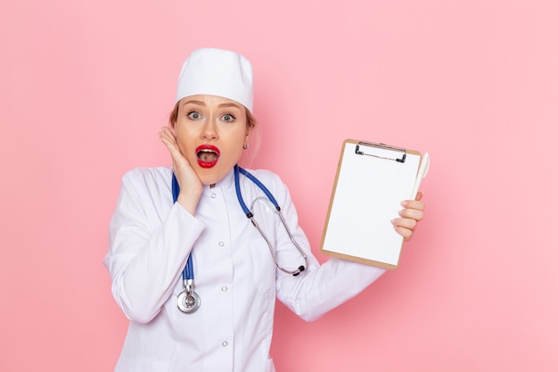 ピンクの宇宙医学病院でポーズをとってメモ帳を保持している青い聴診器で白い医療スーツの正面若い女医