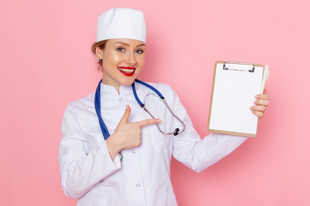 Front view young female doctor in white medical suit with blue stethoscope holding notepad with smile posing on the pink space medicine medical hospital
