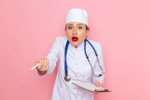 Front view young female doctor in white medical suit with blue stethoscope holding notepad writing down notes on the pink space  job