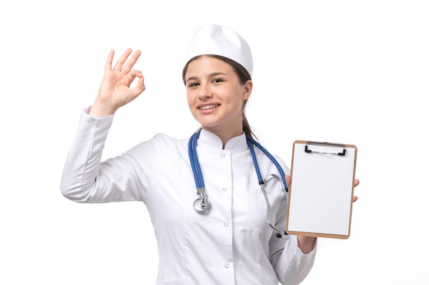 Вид спереди молодая женщина-врач в белом медицинском костюме с голубой стетоскоп, держа блокнот