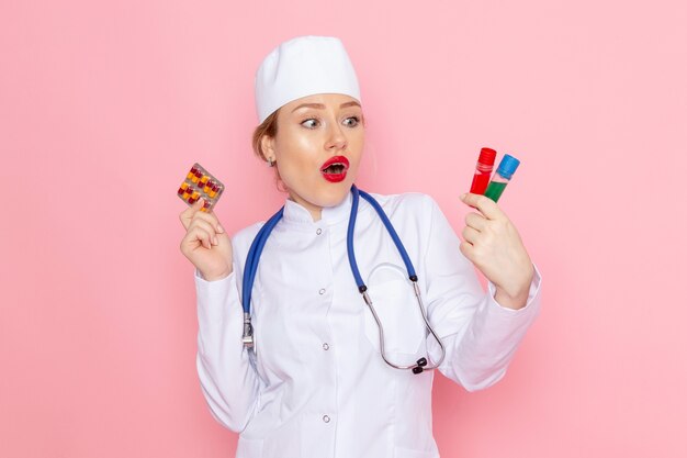 Вид спереди молодая женщина-врач в белом медицинском костюме с синим стетоскопом, держащая таблетки и фляги в медицинской больнице розовой космической медицины