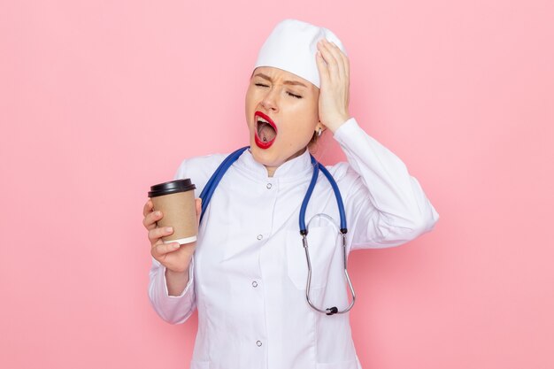 ピンクの宇宙医学病院でプラスチック製のコーヒーカップを保持している青い聴診器で白い医療スーツの正面若い女医