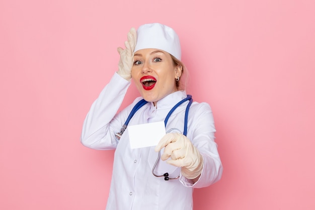 Giovane medico femminile di vista frontale in vestito medico bianco con lo stetoscopio blu che tiene scheda bianca sullo spazio rosa