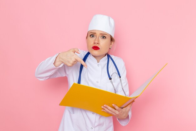 Вид спереди молодая женщина-врач в белом медицинском костюме с синим стетоскопом, держащая желтые файлы на розовой космической работе