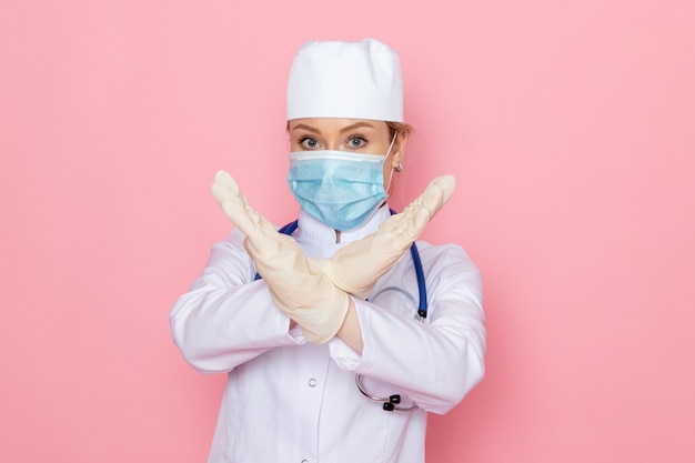 Вид спереди молодая женщина-врач в белом медицинском костюме с синим стетоскопом в стерильной маске на розовой медсестре космической медицины