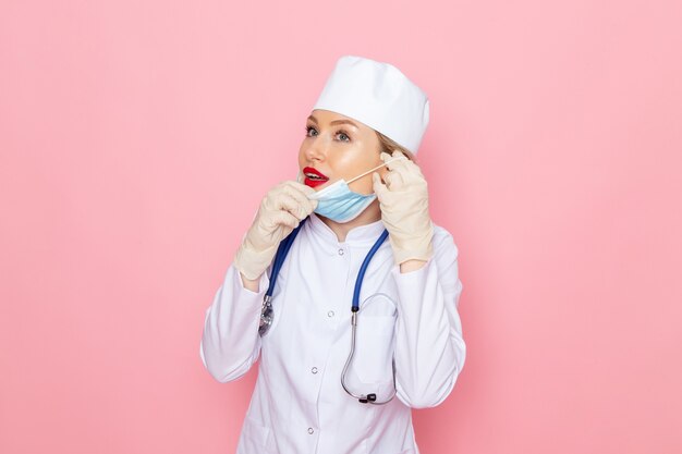 Front view young female doctor in white medical suit with blue stethoscope wearing sterile mask on the pink space 