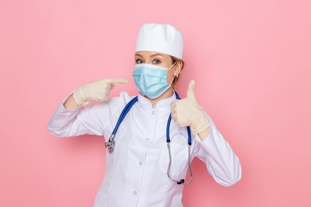 Front view young female doctor in white medical suit with blue stethoscope wearing sterile mask posing on the pink space medical hospital job nurse