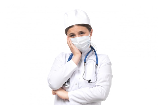 A front view young female doctor in white medical suit with blue stethoscope wearing white mask 