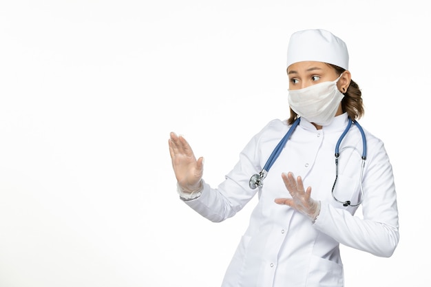 흰색 의료 양복과 밝은 흰색 표면에 코로나 바이러스로 인한 마스크 전면보기 젊은 여성 의사