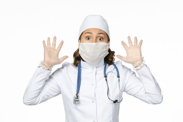 Front view young female doctor in white medical suit and with mask due to coronavirus on light white surface