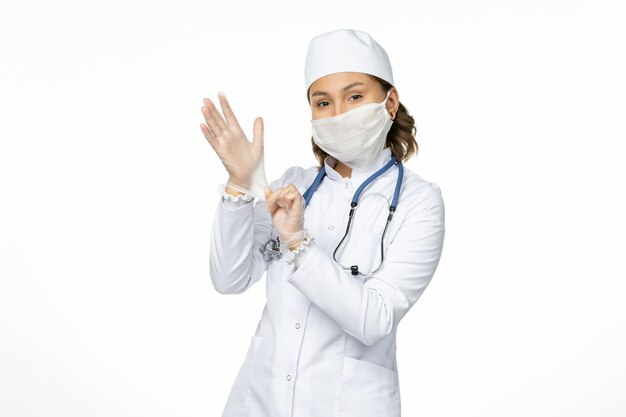 Front view young female doctor in white medical suit and with mask due to coronavirus on the white desk