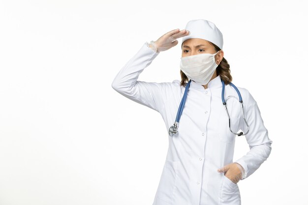 Front view young female doctor in white medical suit and with mask due to coronavirus on white desk