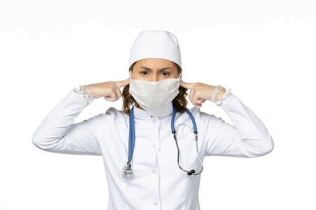 Вид спереди молодая женщина-врач в белом медицинском костюме и с маской из-за коронавируса на белом столе
