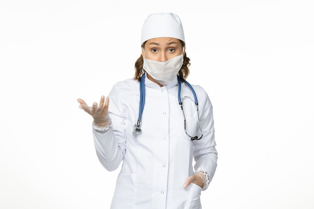Вид спереди молодая женщина-врач в белом медицинском костюме и с маской из-за коронавируса на белой поверхности