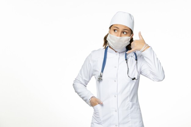 Вид спереди молодая женщина-врач в белом медицинском костюме и стерильной маске из-за коронавируса на светлой белой поверхности