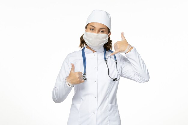 Front view young female doctor in white medical suit and with sterile mask due to coronavirus on white desk