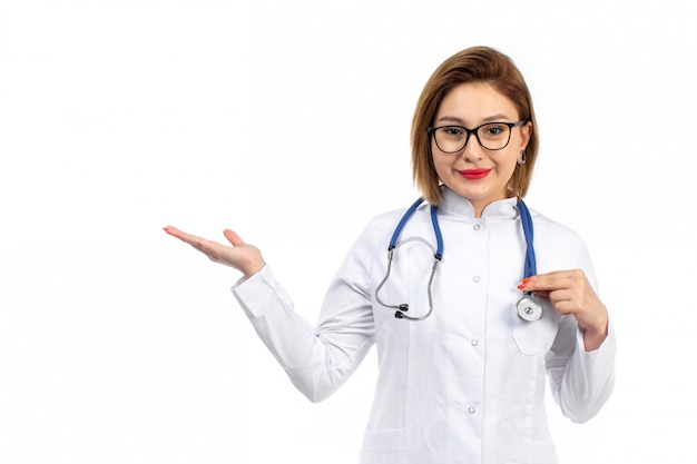 Un giovane medico femminile di vista frontale in vestito medico bianco con lo stetoscopio che sorride sul bianco