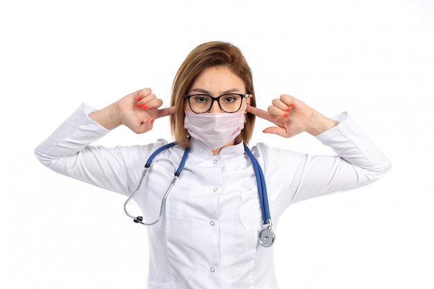 A front view young female doctor in white medical suit with stethoscope wearing white protective mask closing her ears on the white