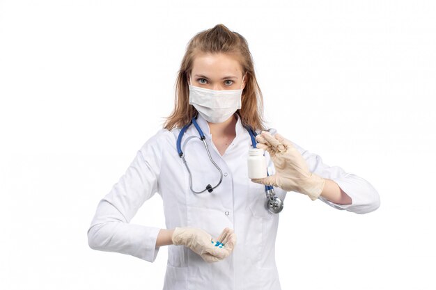 白に薬を保持している手袋で白い防護マスクを着て聴診器で白い医療スーツの正面の若い女性医師