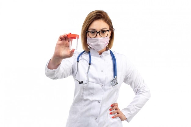 A front view young female doctor in white medical suit with stethoscope wearing white protective mask holding flask on the white