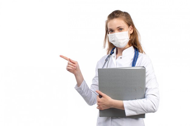 A front view young female doctor in white medical suit with stethoscope wearing white protective mask holding grey files on the white