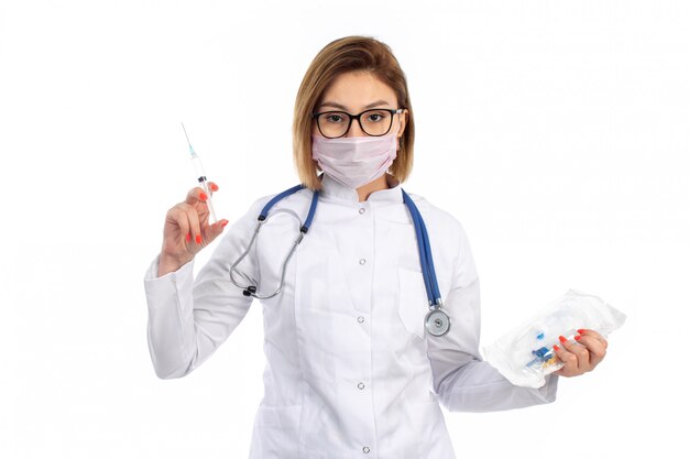 A front view young female doctor in white medical suit with stethoscope wearing white protective mask holding injection on the white