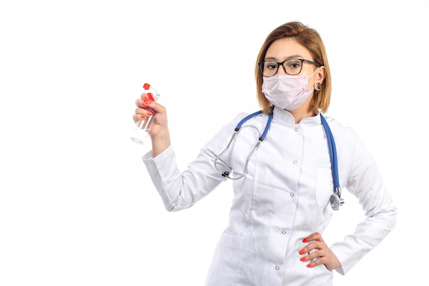 Вид спереди молодая женщина-врач в белом медицинском костюме со стетоскопом в белой защитной маске со спреем на белом фоне