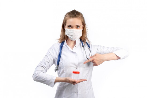 A front view young female doctor in white medical suit with stethoscope wearing white protective mask posing holding flask on the white