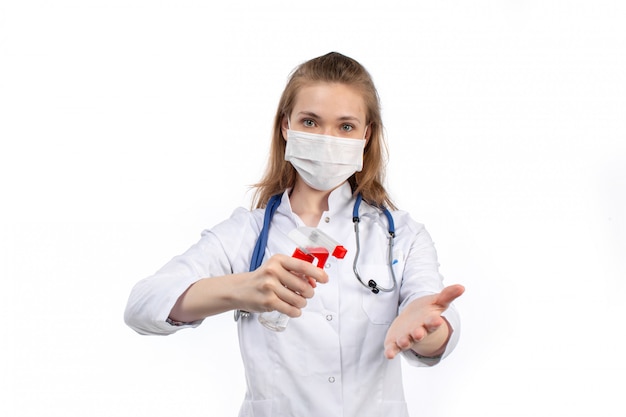 Вид спереди молодая женщина-врач в белом медицинском костюме со стетоскопом в белой защитной маске позирует держа спрей на белом