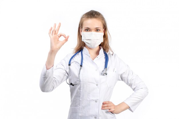 Вид спереди молодая женщина-врач в белом медицинском костюме со стетоскопом в белой защитной маске позирует, показывая хорошо знаком на белом