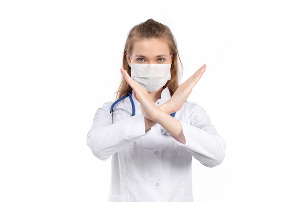 A front view young female doctor in white medical suit with stethoscope wearing white protective mask posing showing ban sign on the white