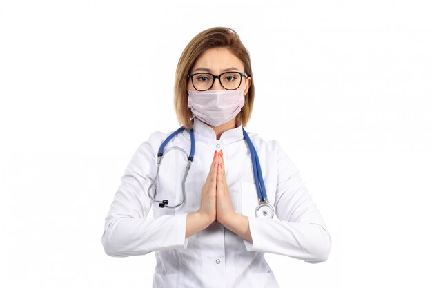 白に祈りのサインを示すポーズをとって白い防護マスクを身に着けている聴診器で白い医療スーツの正面の若い女性医師
