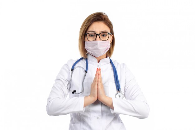 A front view young female doctor in white medical suit with stethoscope wearing white protective mask posing showing pray sign on the white