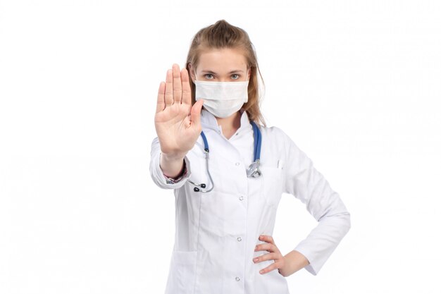 A front view young female doctor in white medical suit with stethoscope wearing white protective mask posing showing stop sign on the white