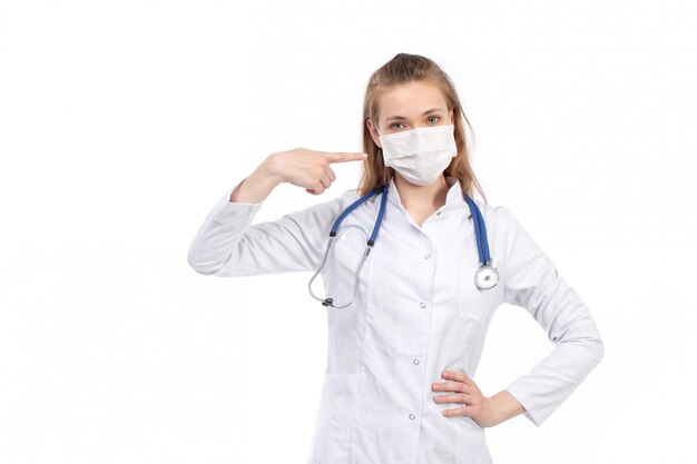 A front view young female doctor in white medical suit with stethoscope wearing white protective mask posing on the white