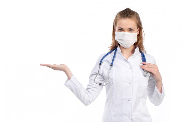 A front view young female doctor in white medical suit with stethoscope wearing white protective mask posing on the white