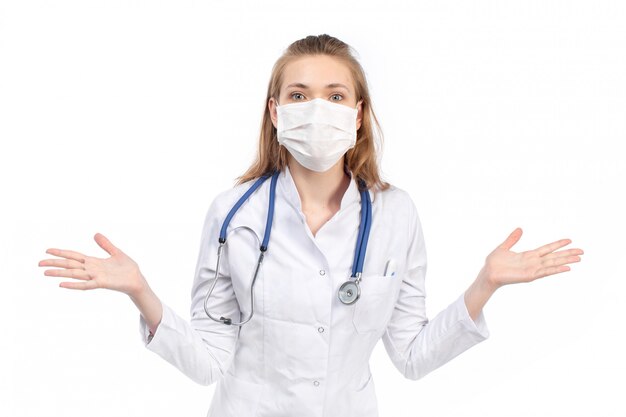 A front view young female doctor in white medical suit with stethoscope wearing white protective mask posing on the white